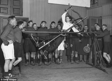 woman demonstrating an exercise machine, known as the Gymo Frame, to members of the Arsenal soccer team at Highbury football ground in London, UK, in 1932. 